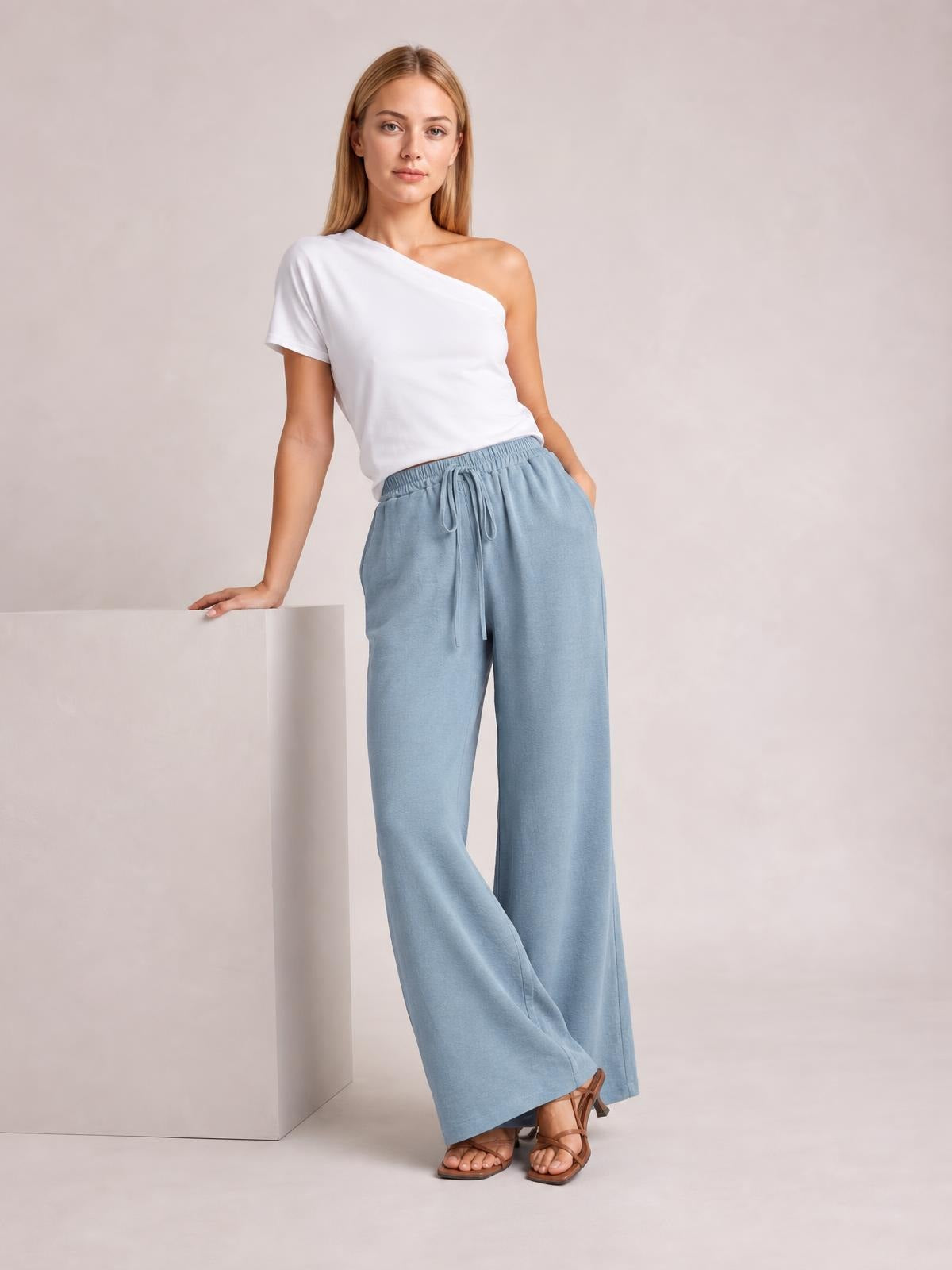 Model wears light blue wide leg drawstring pants with an elasticized waist and flowing full-length silhouette, posing in flat strappy sandals.