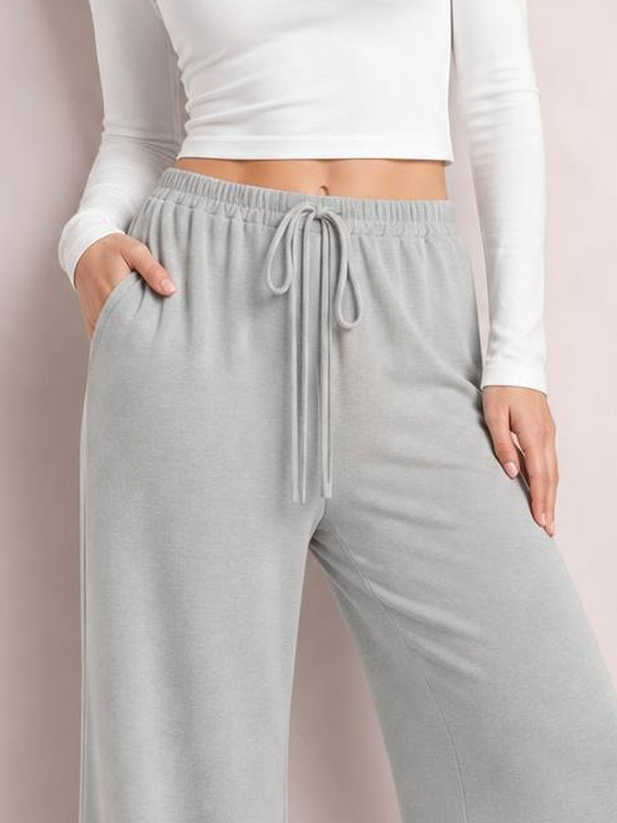 Model wearing light gray wide leg drawstring pants with an elastic waistband, long tie, roomy flowy silhouette, and hands resting in the side pockets.
