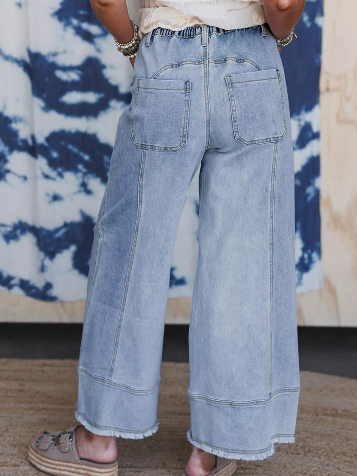 Model stands with hands in pockets wearing light-wash wide-leg drawstring jeans featuring large back patch pockets, seamed panels, and raw hems.