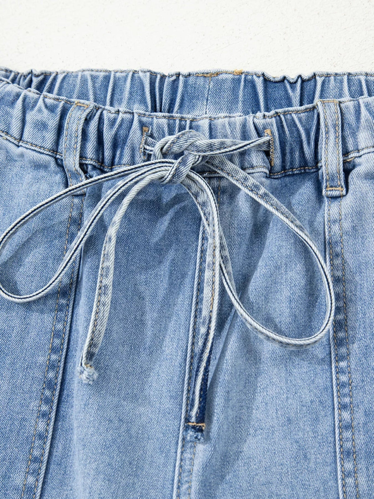 Close-up of light blue wide-leg jeans showing an elasticized drawstring waistband, belt loops, front pockets, and detailed contrast stitching.