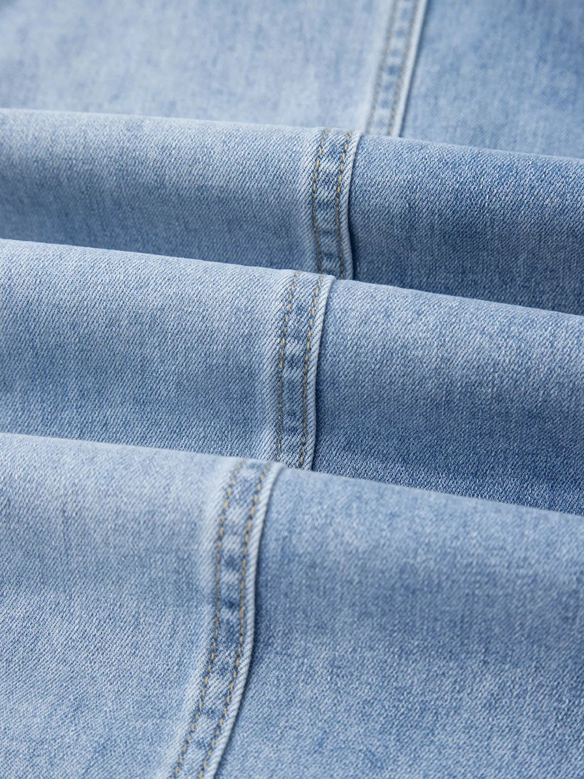 Close-up of light blue denim jeans showing sturdy twill fabric, overlapping panels, and neat double-stitched construction seams.