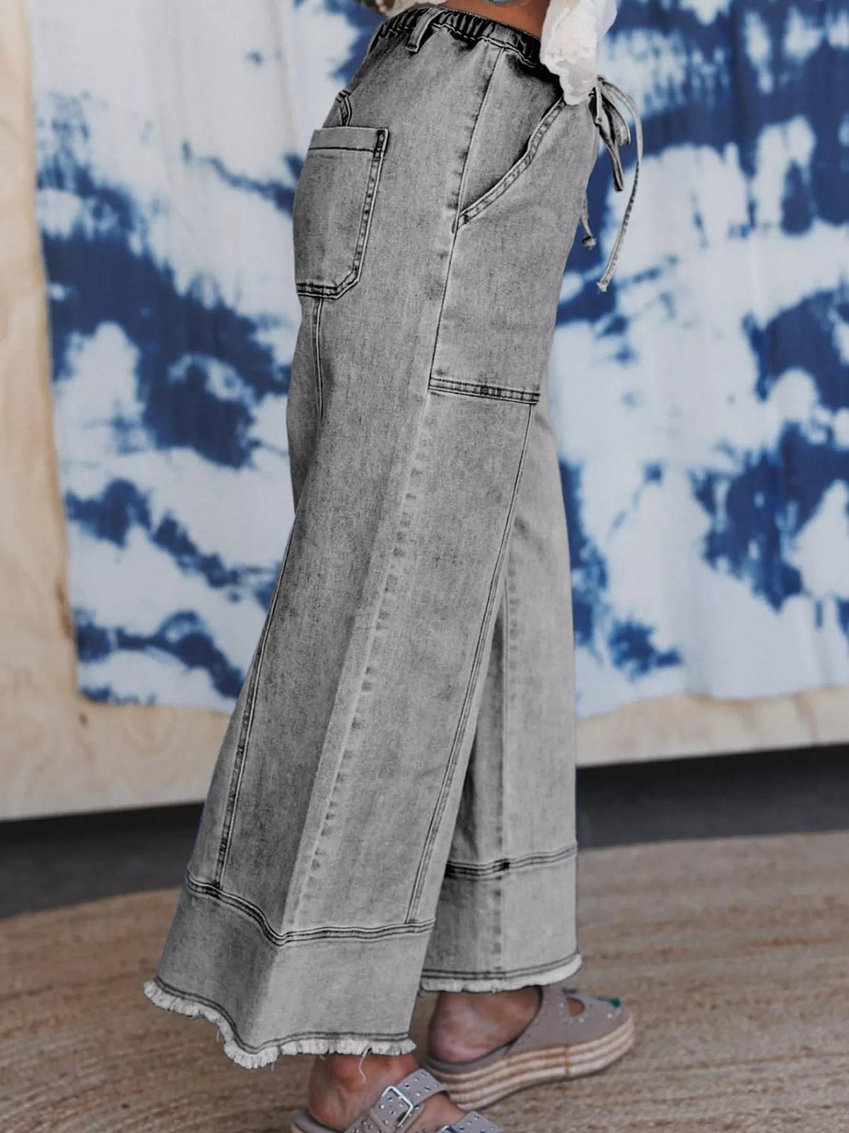 A model wears gray wide-leg drawstring jeans in a relaxed silhouette, featuring large patch pockets, contrast seaming, and raw hems.