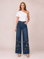 Model wearing dark blue wide-leg jeans with a high-rise waistband, front pockets, straight seams, and gold-tone eyelet details throughout the lower legs.