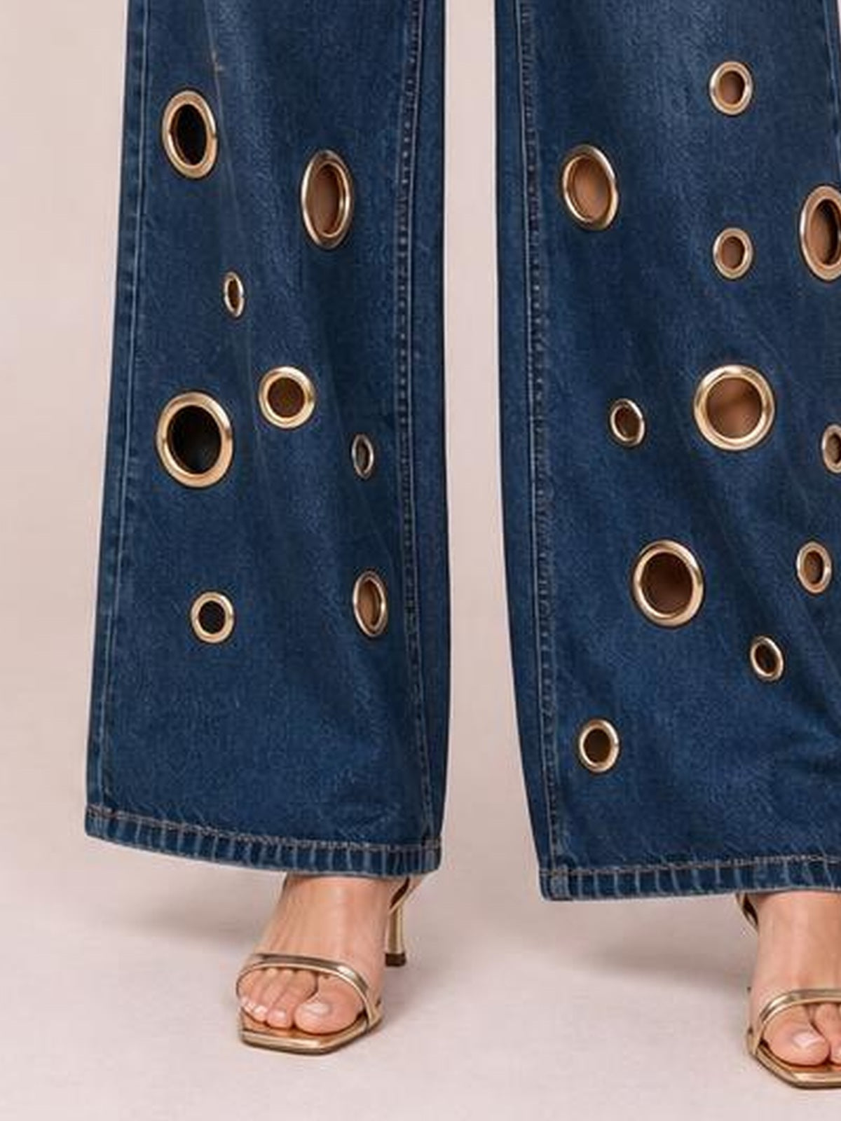 Close-up of blue wide-leg jeans with gold eyelet details, straight stitched construction, and cropped hems shown on a standing model.