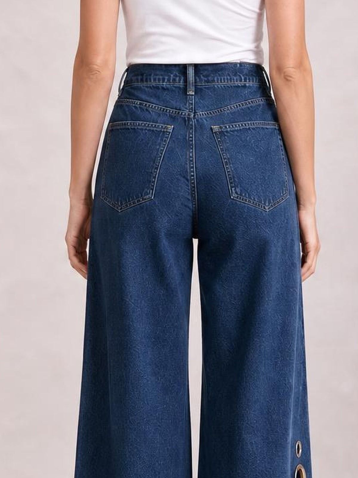 Model standing from behind wears dark blue wide-leg jeans with a structured waistband, contrast stitching, back patch pockets, and eyelet details near the lower outer leg.