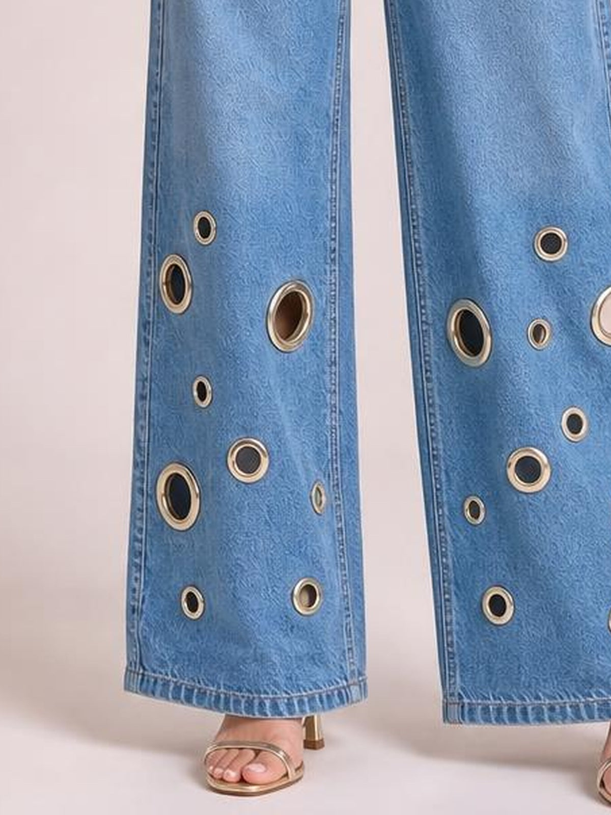 A model wears light blue wide leg jeans with stitched hems and metallic eyelet details arranged along the lower legs, paired with strappy sandals.