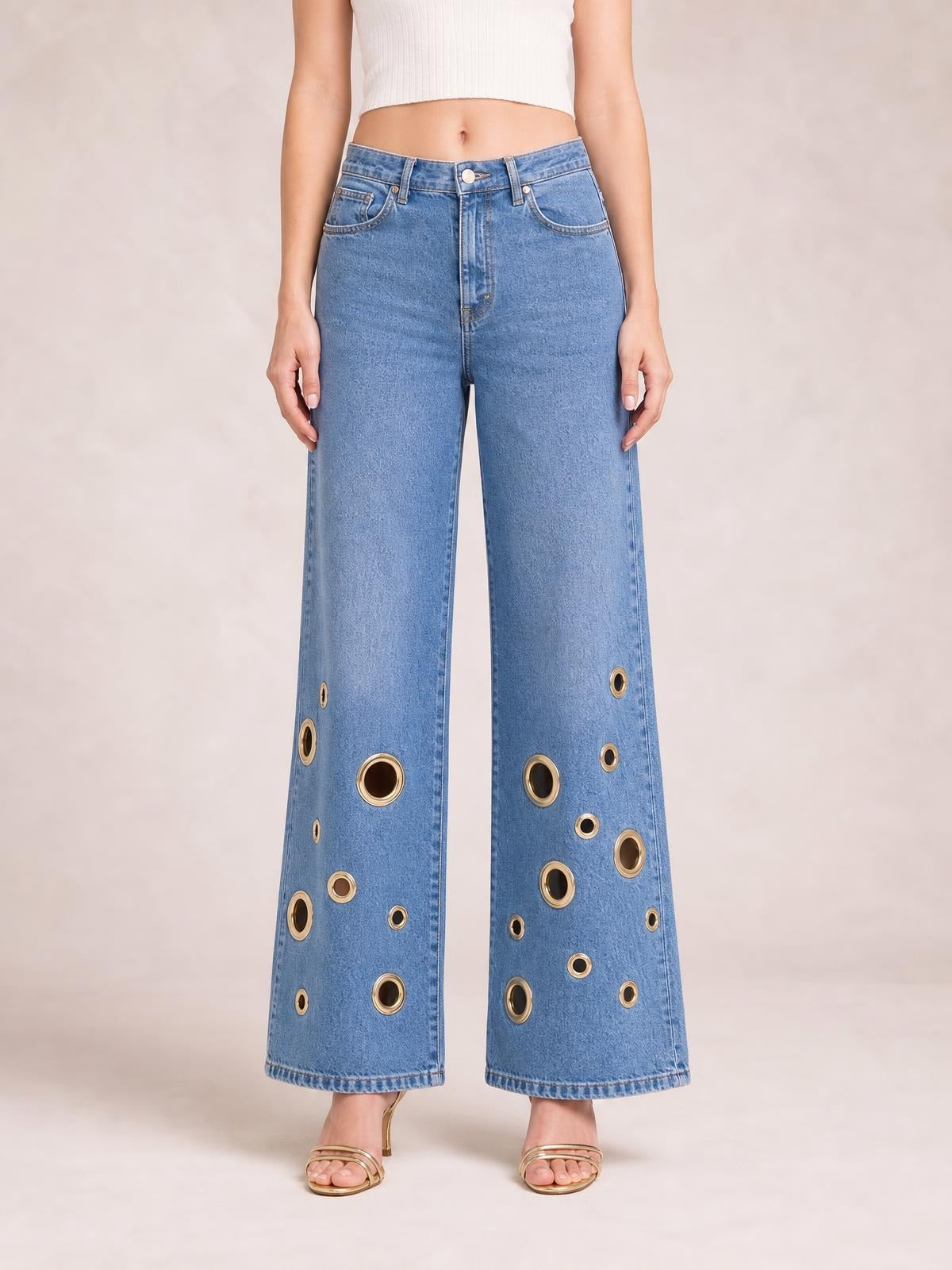 Model stands wearing light blue wide leg jeans with a high waist, five-pocket construction, and multiple gold-tone eyelet details along the lower legs.