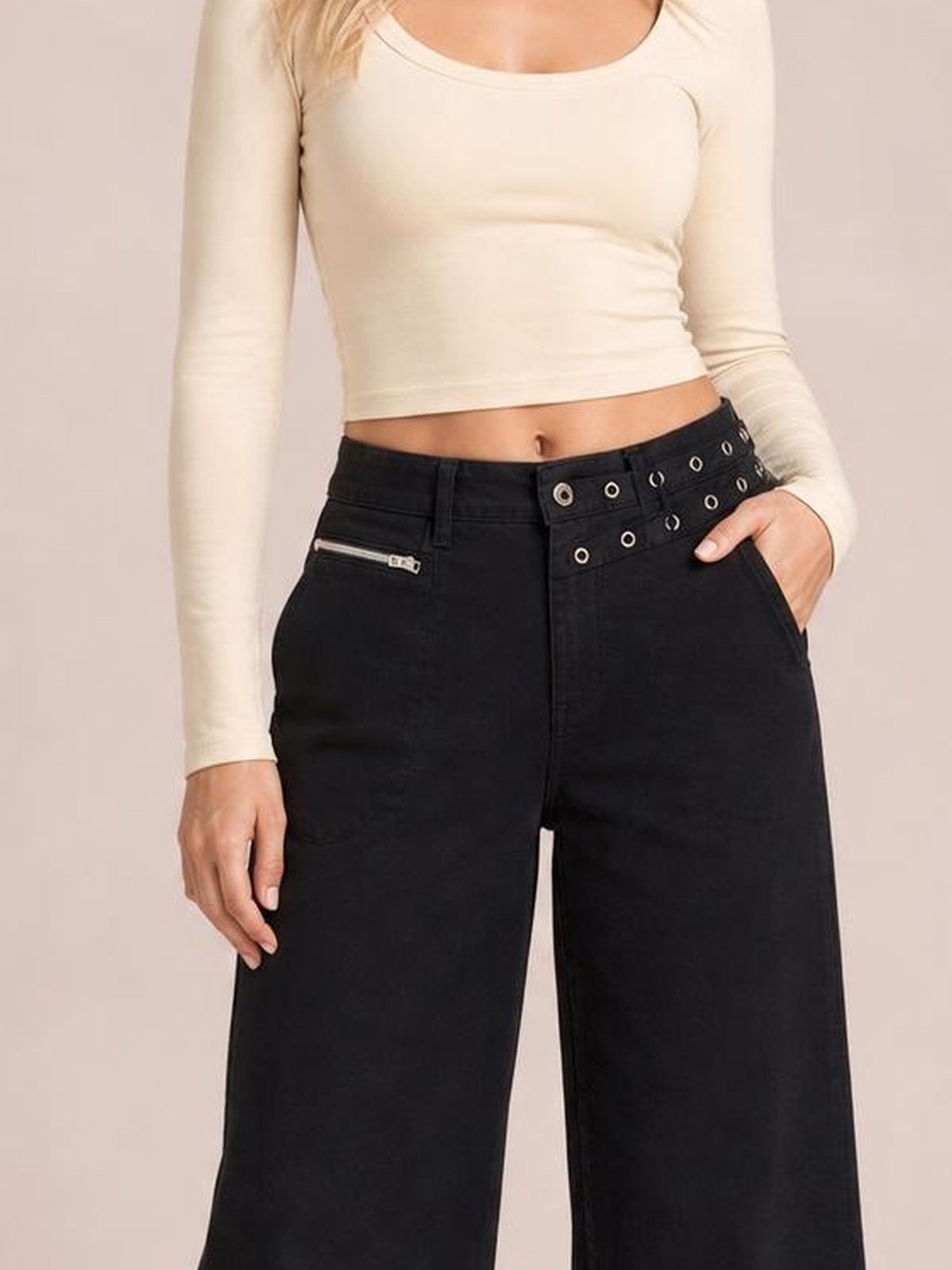 Model wearing black wide leg jeans with front pockets, belt loops, a zippered pocket, and a decorative grommet waistband, posed with one hand in a pocket.
