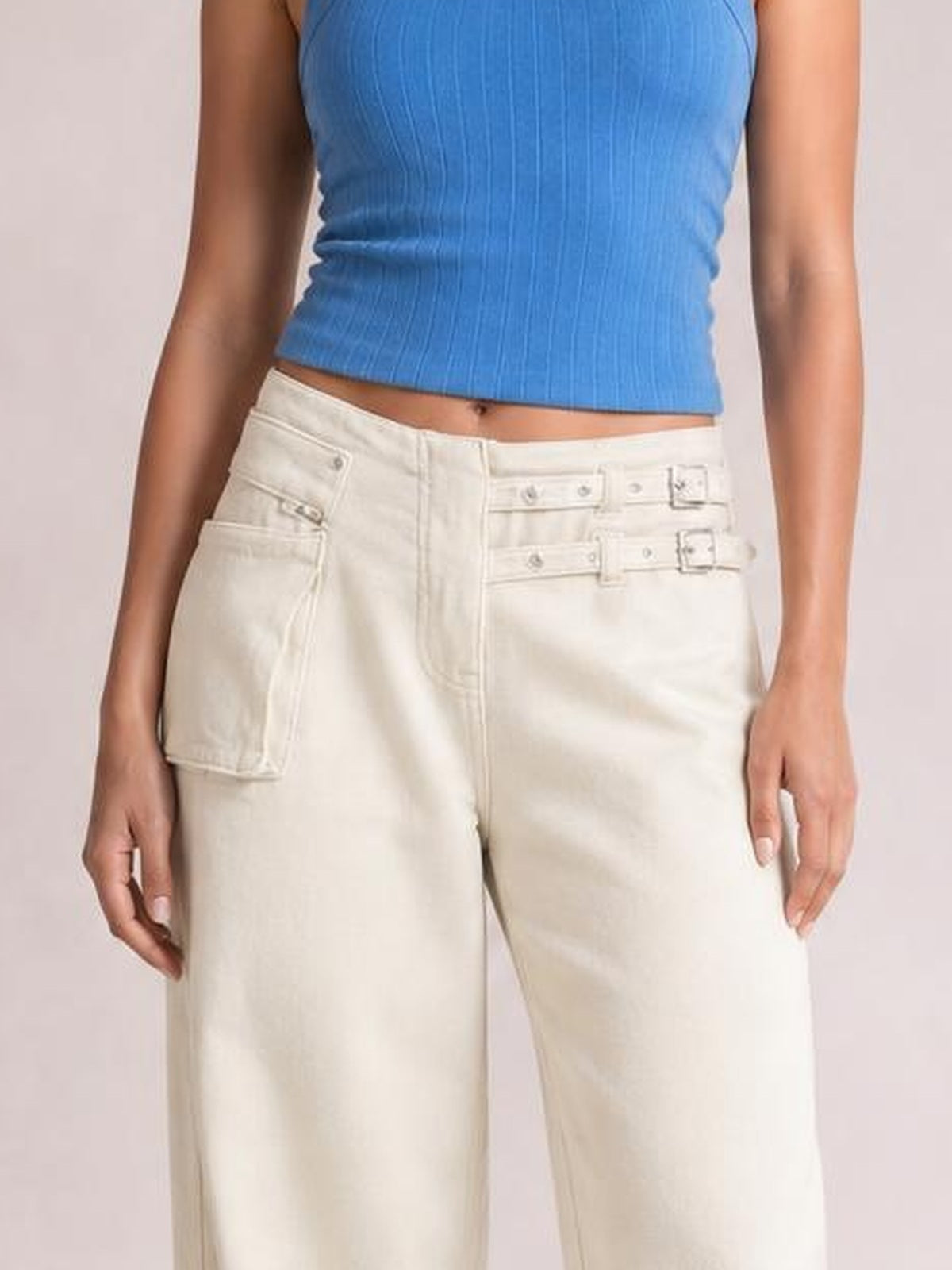 Model wearing cream wide leg jeans with large pockets, multiple belt loops, and flap details, shown with hands resting at her sides.