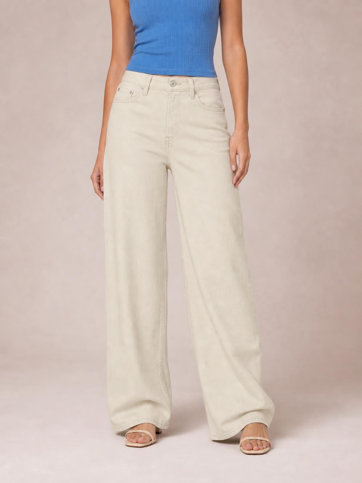 A model stands wearing cream wide leg jeans with visible front pockets, belt loops, button closure, contrast stitching, and a relaxed full-length silhouette.