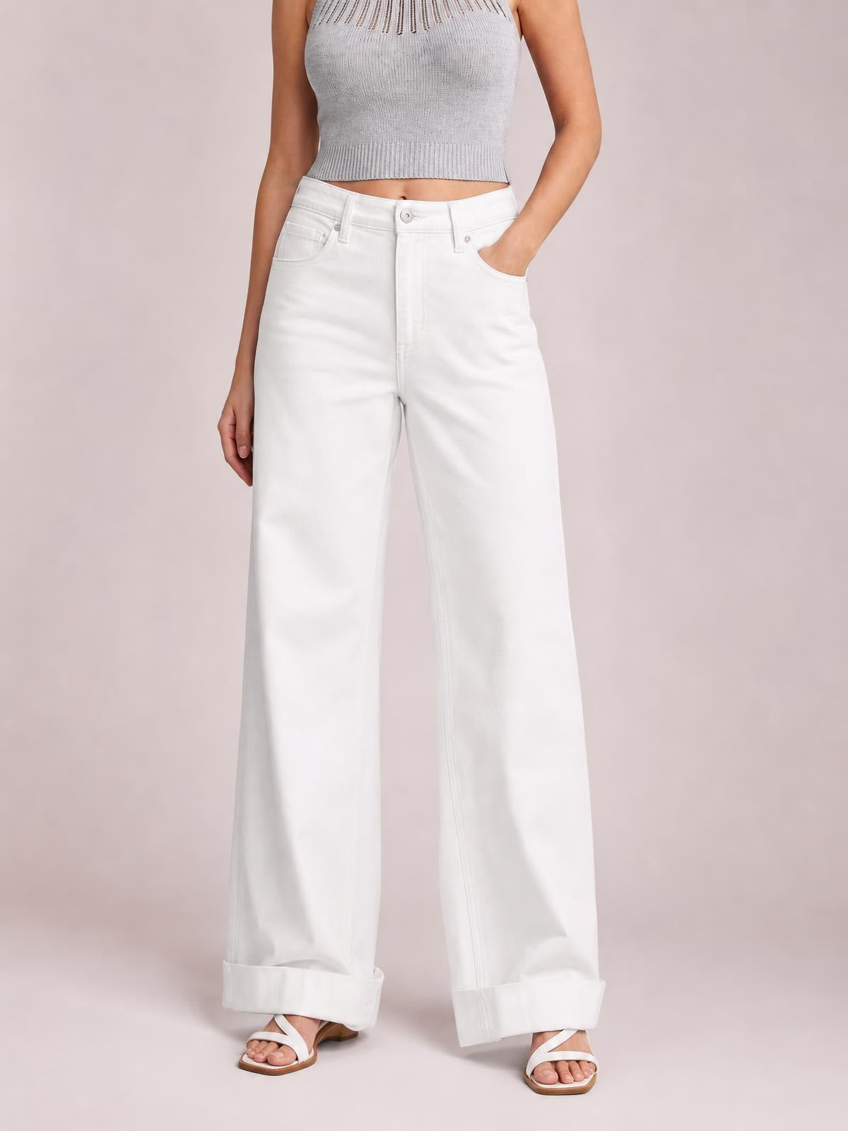 Standing model wears white wide-leg jeans with a button closure, front pockets, straight construction, and rolled cuffs, shown with one hand in a pocket.