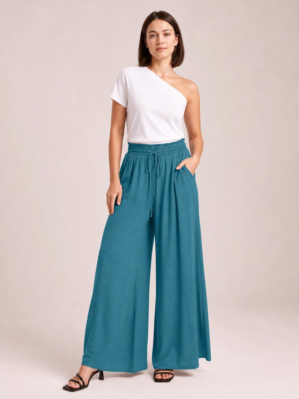 Model wearing teal wide leg pants with pockets, a gathered elastic waistband, and front drawstring, styled with a white one-shoulder top.