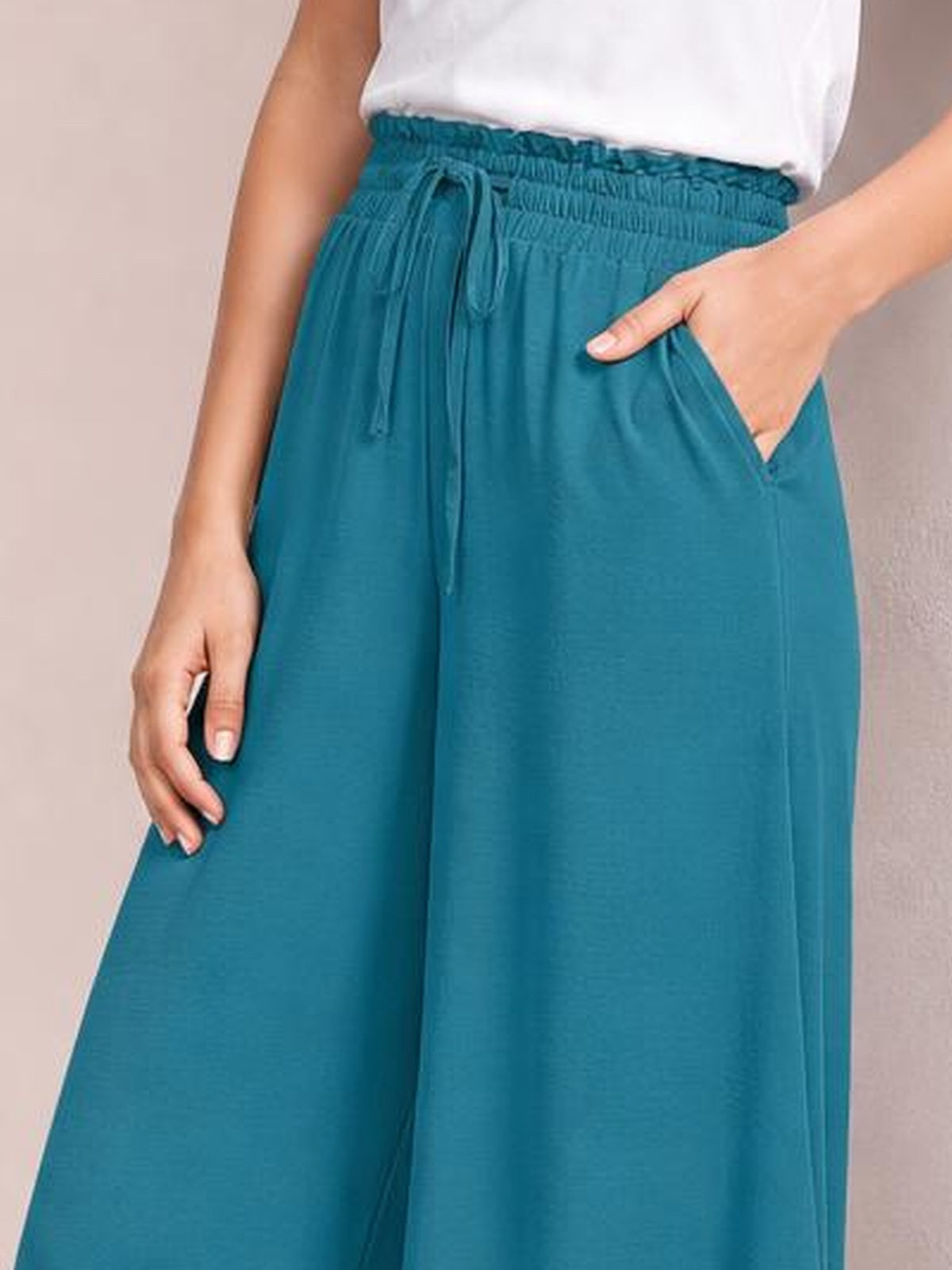 A model wears teal wide leg pants with an elastic waistband, front drawstring, and practical side pockets, shown with one hand tucked into a pocket.
