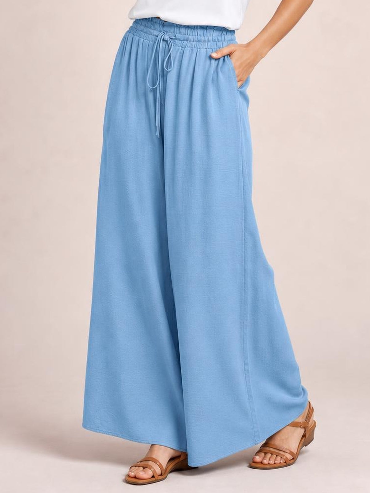 Model wearing light blue wide leg pants with a drawstring waistband and side pockets, standing in brown sandals with one hand tucked into a pocket.