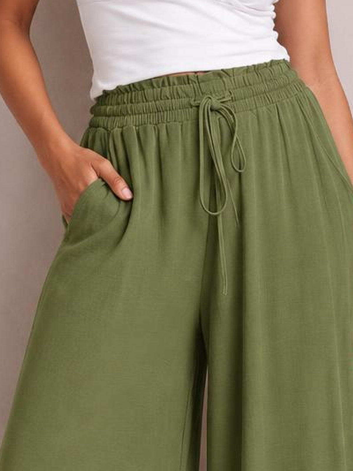 A cropped model wears olive green wide leg pants with an elastic drawstring waist and practical side pockets, shown with one hand tucked into a pocket.