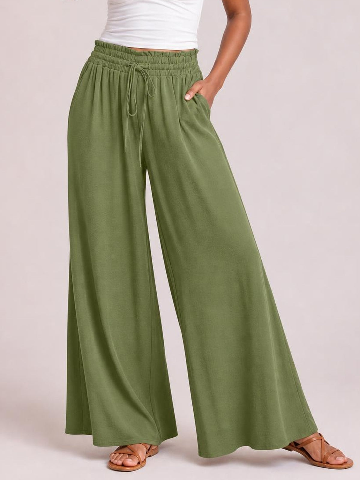 A model wears olive green wide leg pants with a relaxed silhouette, gathered drawstring waist, flowing legs, and a side pocket, posing with one hand tucked inside.