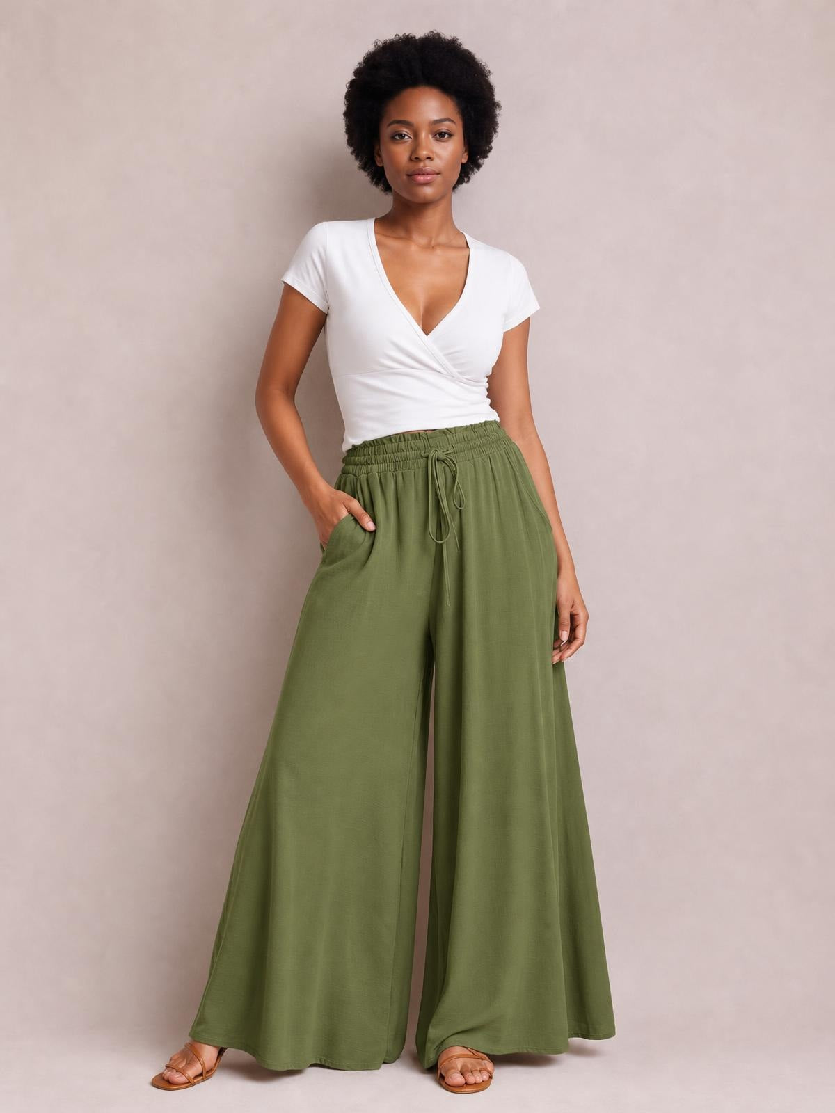 Model wearing olive green wide leg pants with side pockets, an elastic drawstring waist, and a flowing floor-length silhouette, posing with one hand in a pocket.