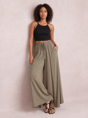 Model poses in taupe wide leg pants with an elastic drawstring waist and side pockets, one hand tucked into a pocket and legs crossed.