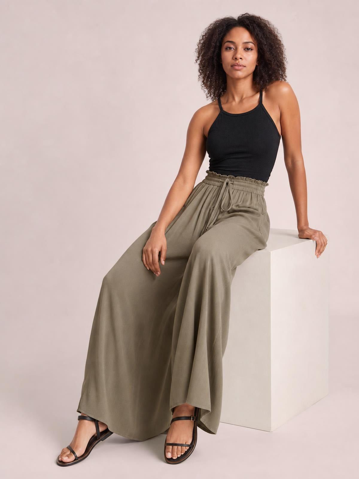 A model sits in taupe wide leg pants with a relaxed flowing silhouette, elastic drawstring waist, and practical pockets, styled with a black camisole.