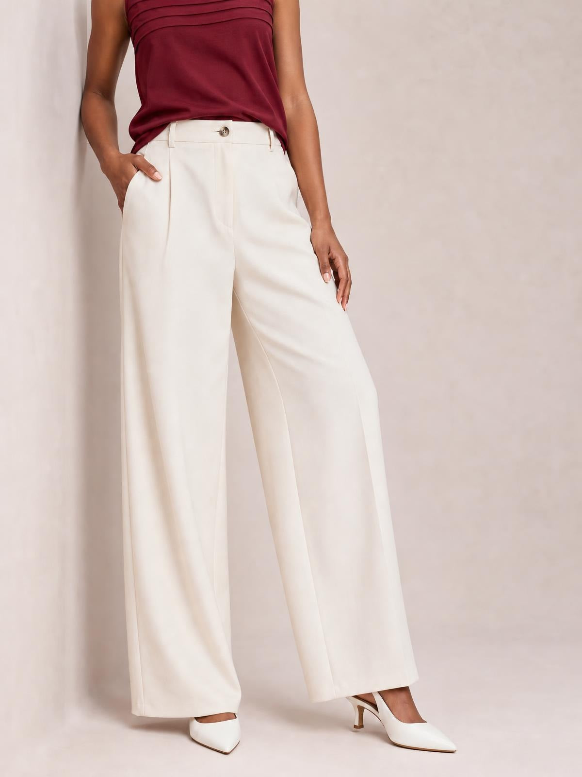 Model wearing white wide-leg pleated pants with a buttoned waistband, front pleats, and long flowing legs, posing with one hand in a pocket.