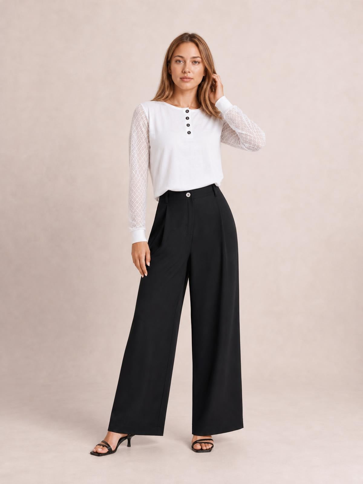 A standing model wears black wide-leg pleated pants with a buttoned waistband and flowing full-length silhouette, posed with one hand at her hair.