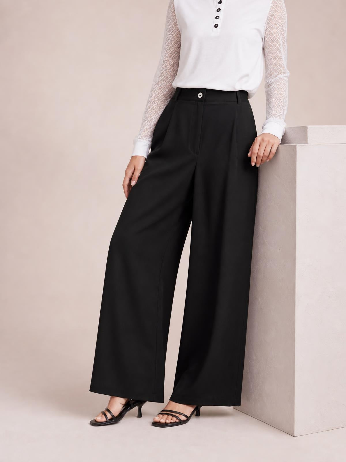 A model stands in black wide-leg pleated pants with a buttoned waistband, belt loops, front pleats, and a fluid full-length silhouette.