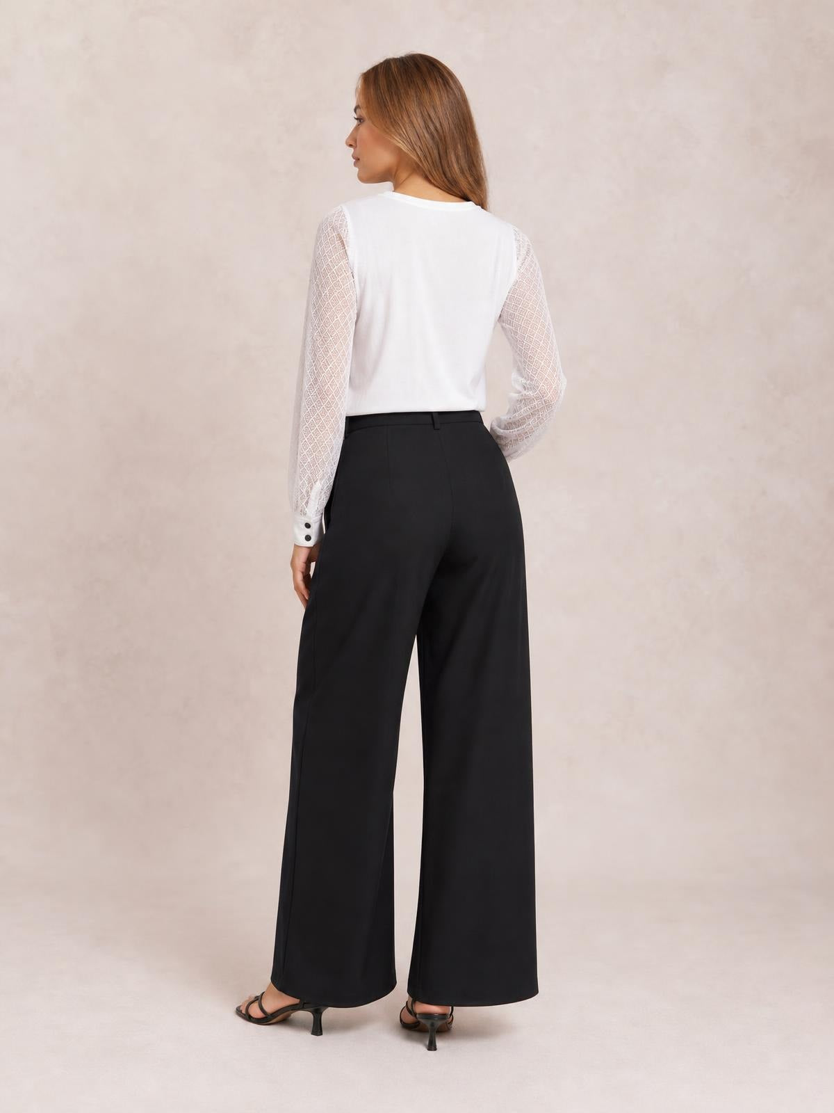 A model looks over her shoulder in black wide-leg pleated pants with a high waistband, belt loops, and a flowing full-length silhouette.