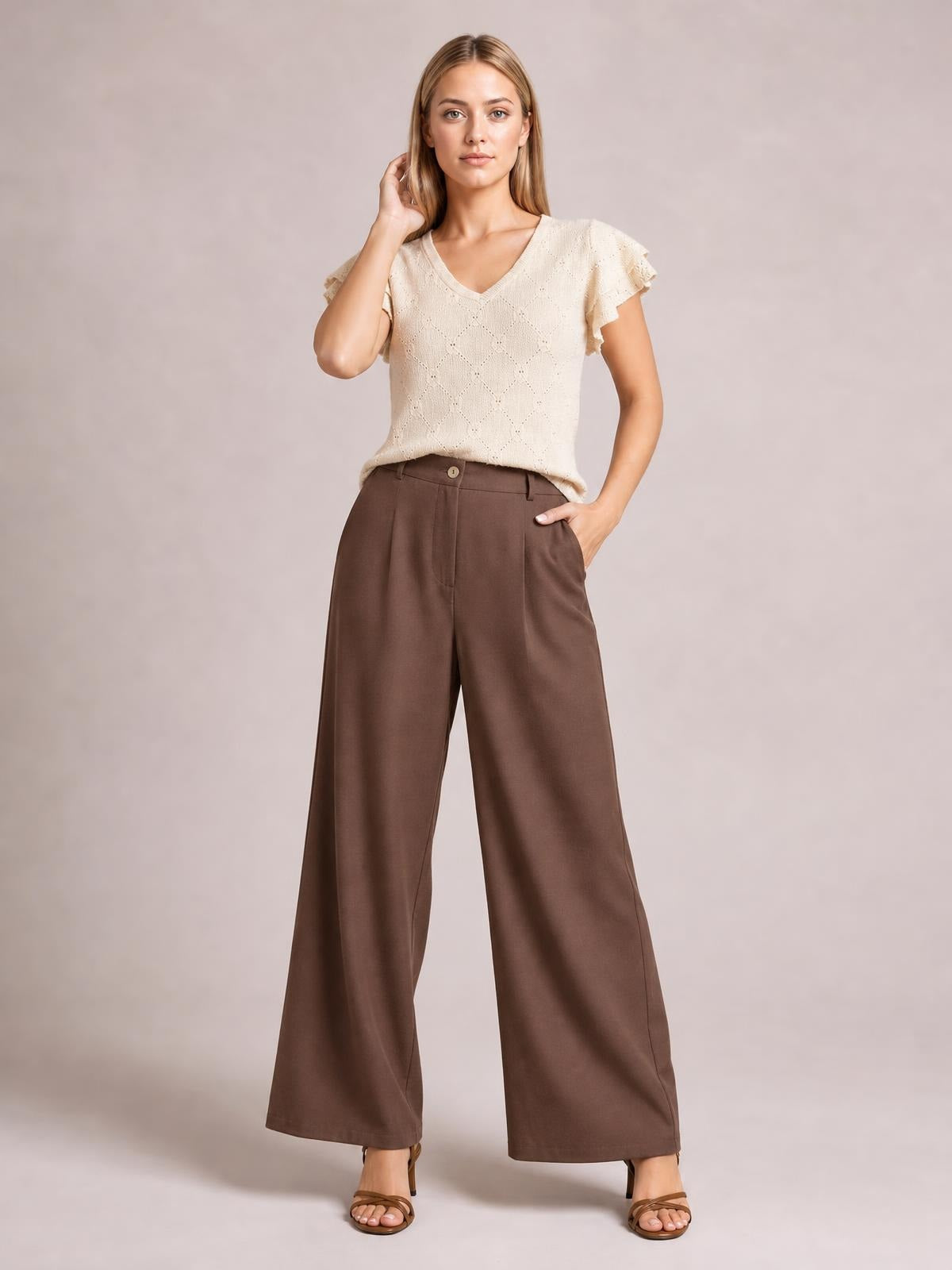 Model wears brown wide leg pleated pants with a buttoned high waist, front pleats, belt loops, and a flowing full-length silhouette.