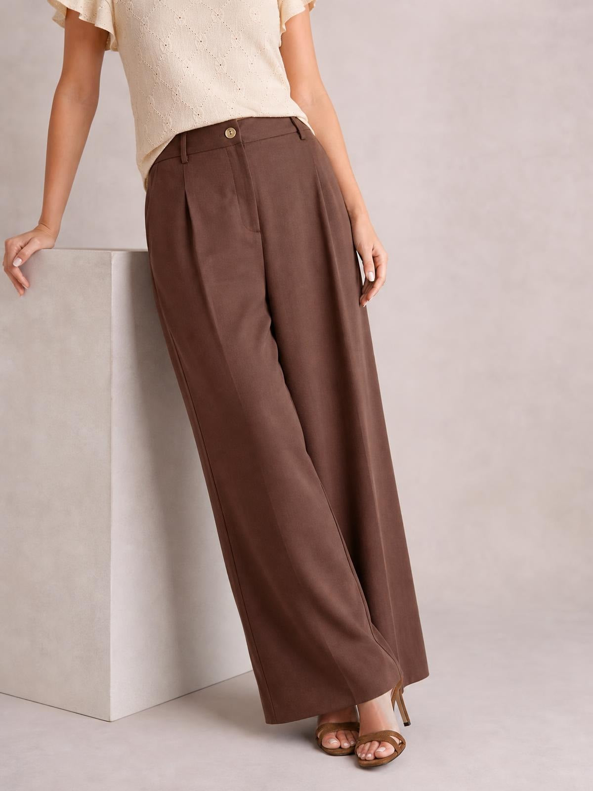 A model leans casually while wearing brown wide leg pleated pants with a buttoned waistband, front pleats, and a flowing full-length silhouette.