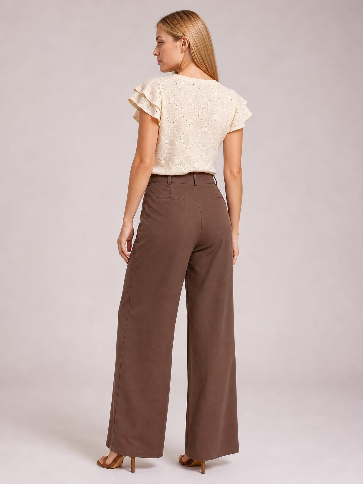 A model shown from behind wears brown wide-leg pleated pants with a tailored waistband, belt loops, back pockets, and a long flowing silhouette.