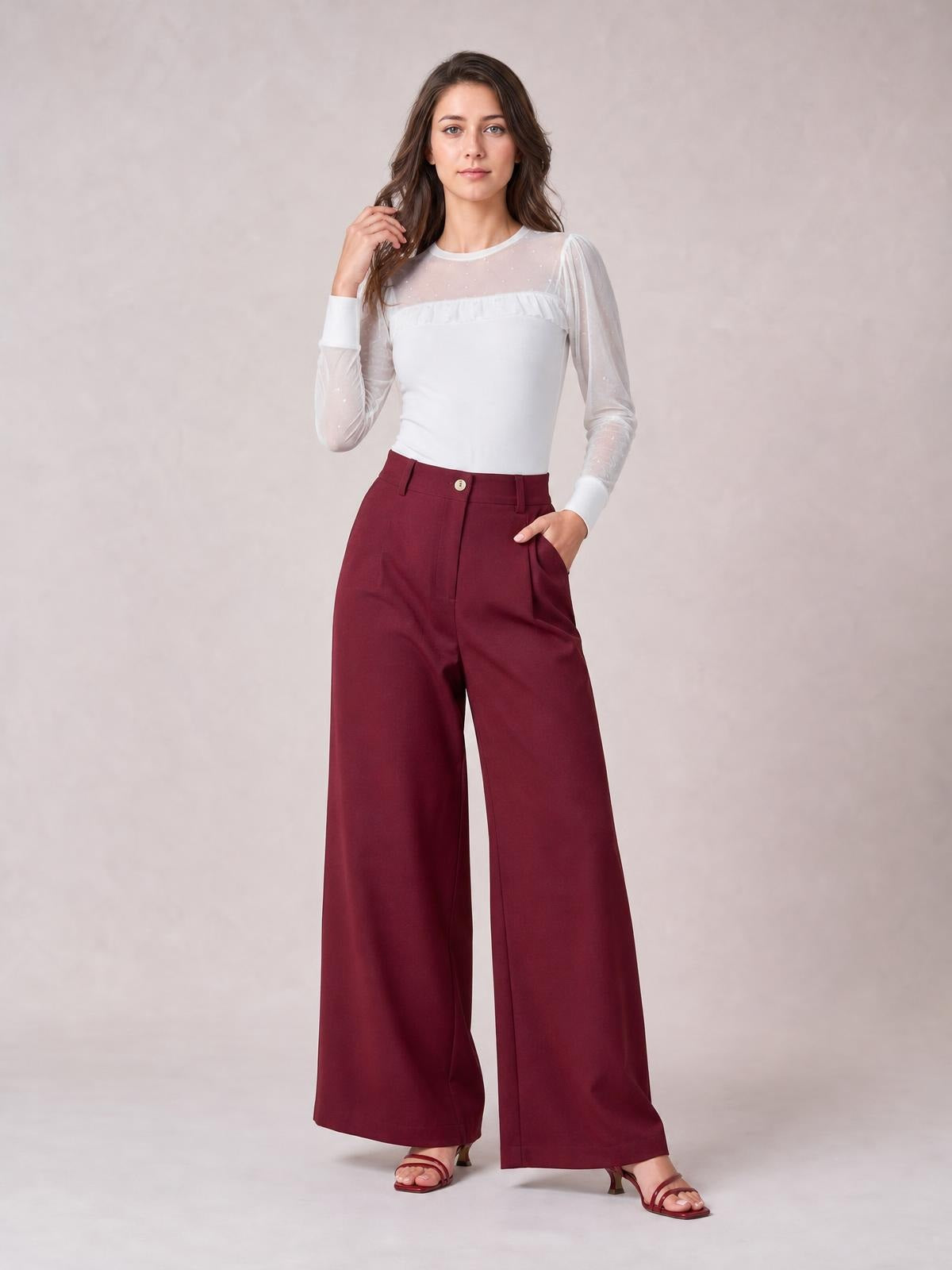 A model poses in burgundy wide-leg pleated pants with a high waist, button closure, belt loops, side pockets, and a fluid floor-length silhouette.