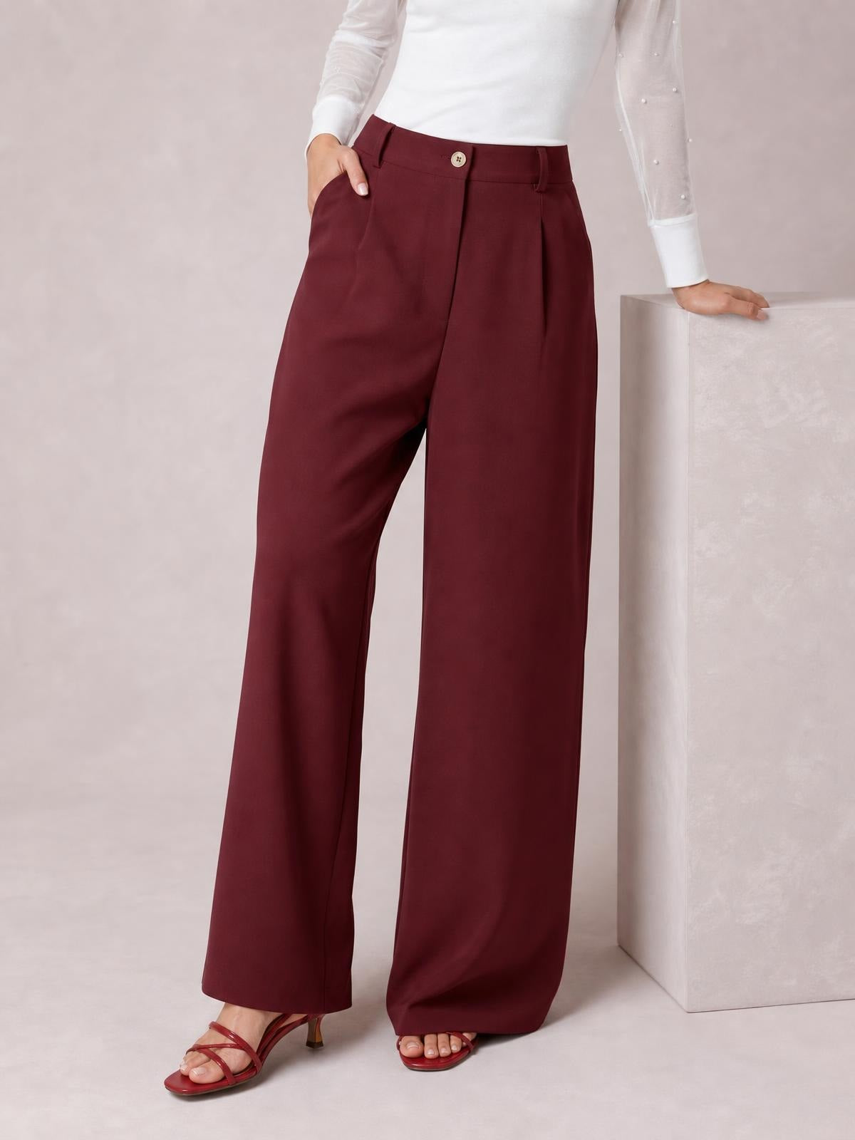 Model wearing burgundy wide leg pleated pants with a buttoned front, belt loops, slanted pockets, and a flowing full-length silhouette.
