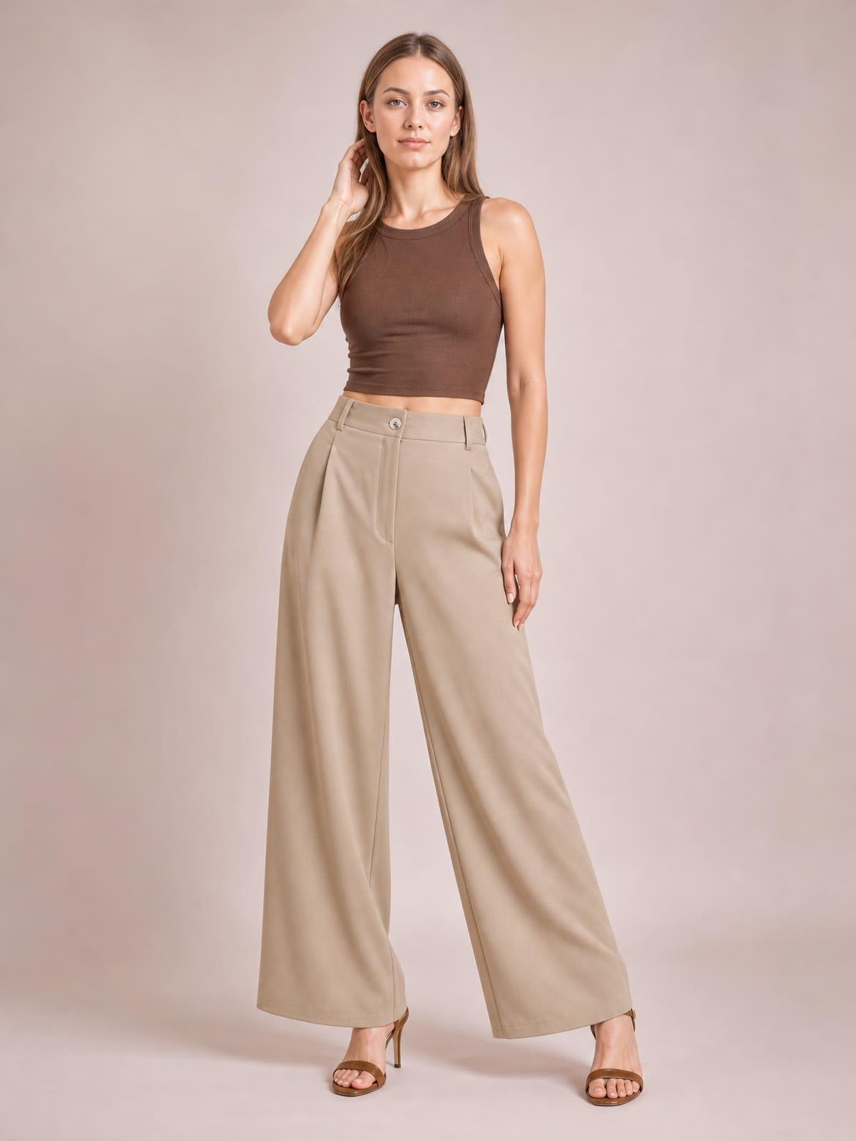 A model stands with one hand touching her hair, wearing beige wide-leg pleated pants with a buttoned waistband, belt loops, and flowing full-length legs.