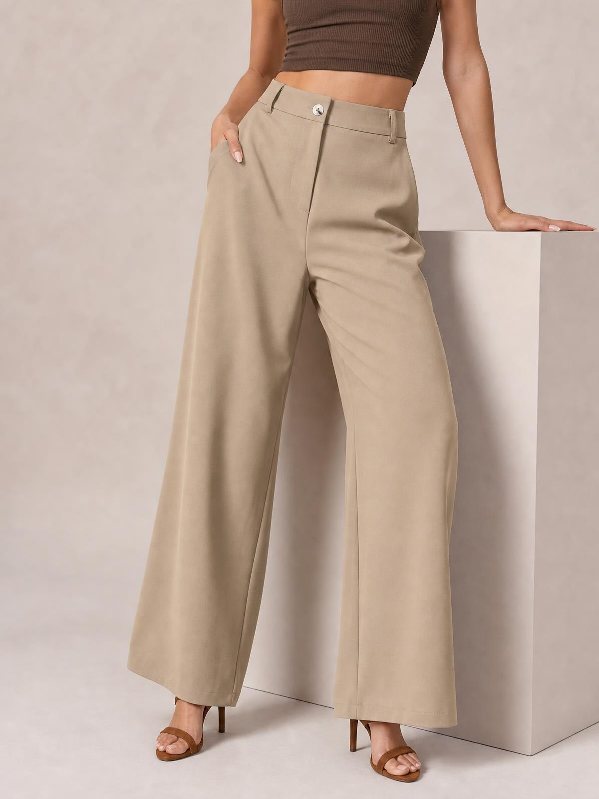 Model wearing beige wide-leg pleated pants with a buttoned high waistband, belt loops, front pockets, and a relaxed full-length silhouette.