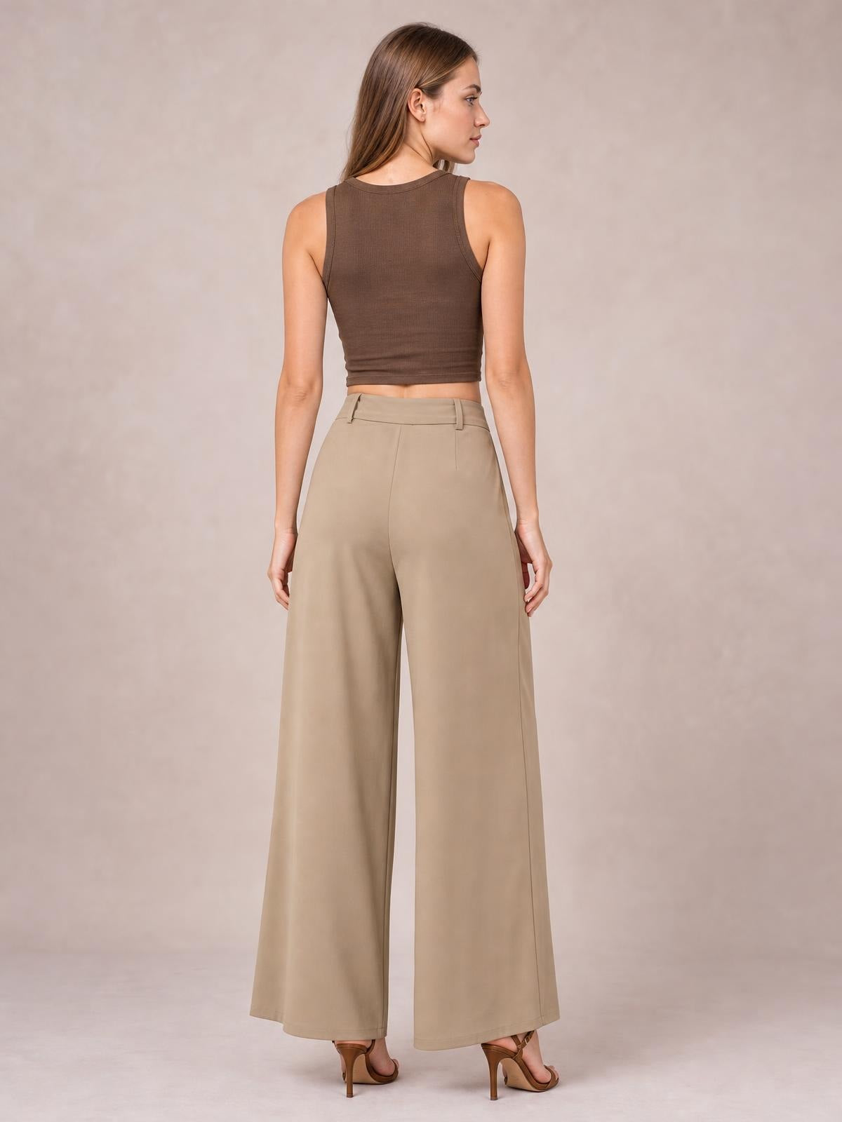 Model shown from the back wearing beige wide-leg pleated pants with a high waist, belt loops, button closure, and full-length flowing silhouette.