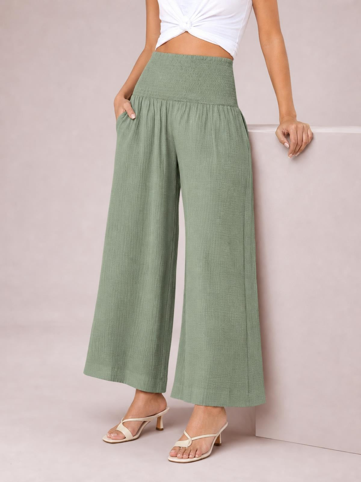 Model wearing sage green wide-leg pants with a high smocked waist, gathered construction, and flowing full-length silhouette, posing with one hand in a pocket.
