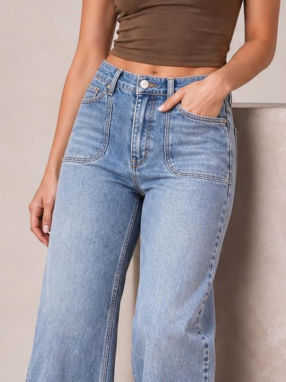 A model poses with one hand in the pocket, showcasing light blue wide-leg wash jeans with a button fly, belt loops, curved front pockets, and contrast stitching.