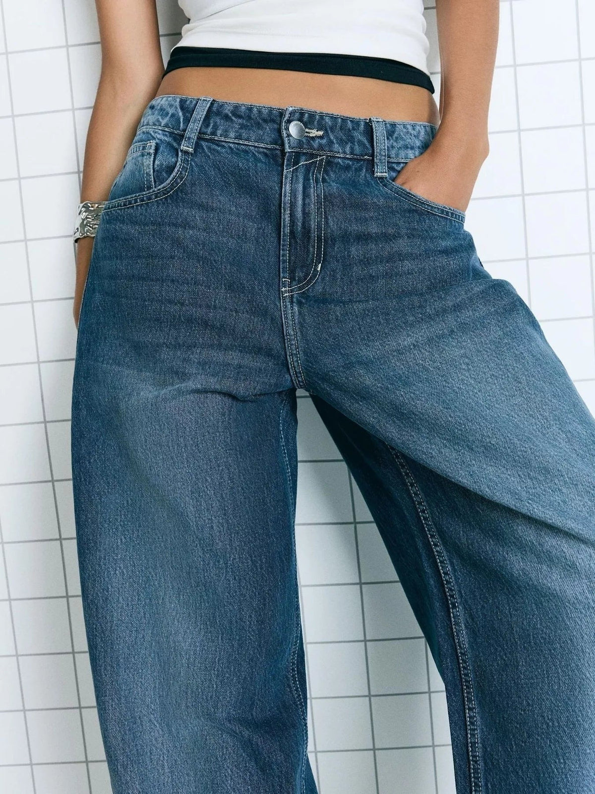 A model wears blue washed wide leg jeans with a buttoned waistband, belt loops, front pockets, contrast stitching, and a relaxed silhouette.