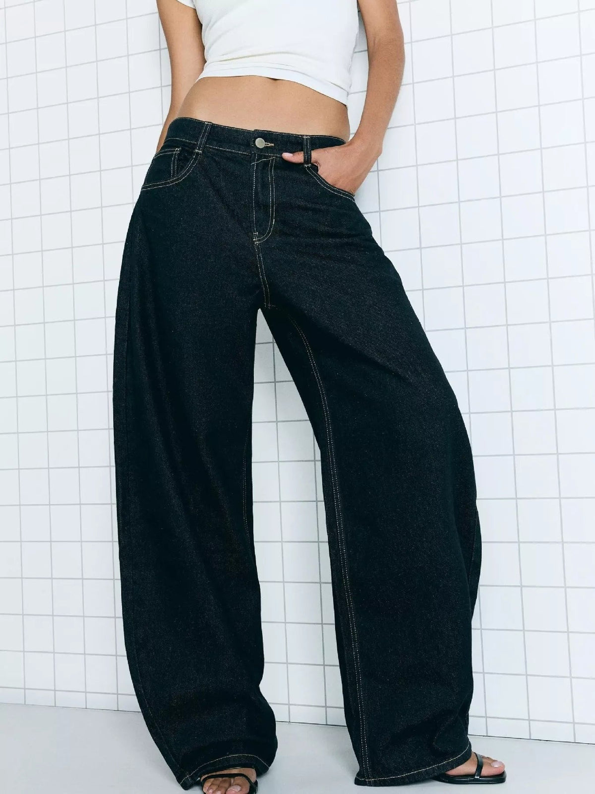 Model wearing dark washed wide-leg jeans with contrast stitching, button closure, front pockets, and a relaxed full-length silhouette, one hand in a pocket.