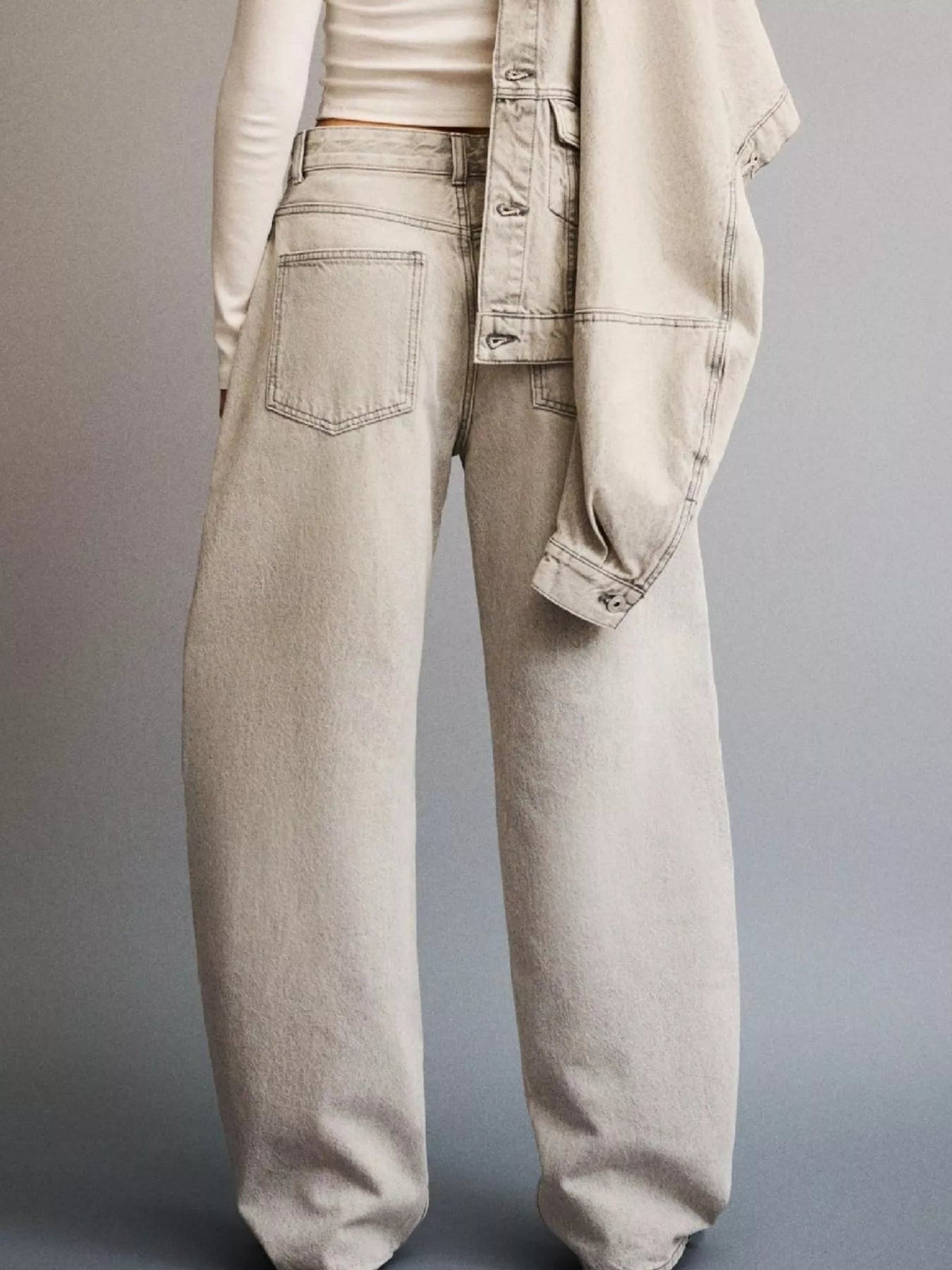 Model wears light washed wide leg jeans with a relaxed silhouette, button closure, belt loops, and visible back pockets while holding a denim jacket at the hip.