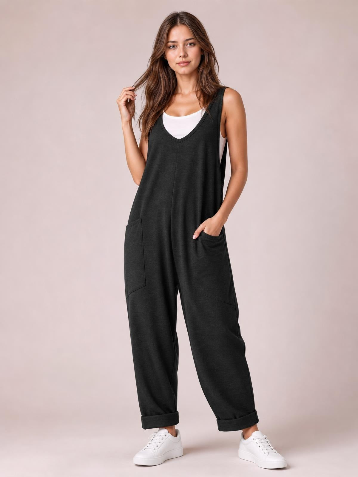 Model poses in a black wide-strap jumpsuit with a V-neckline, relaxed wide-leg silhouette, cuffed hems, and large front pockets.
