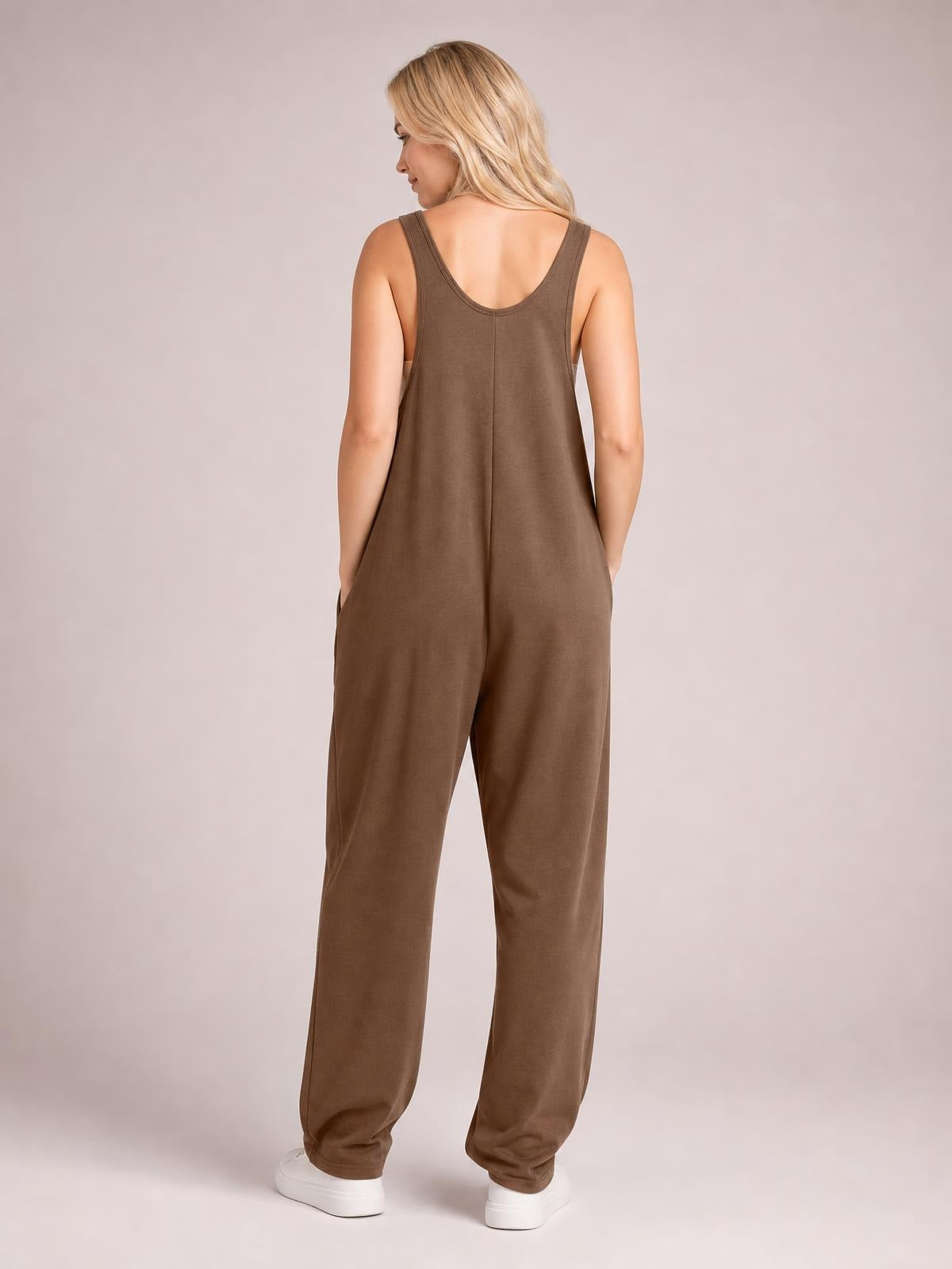 Model turned away in a brown wide-strap jumpsuit with a relaxed, loose-leg silhouette, scoop back, center seam, and side pockets.