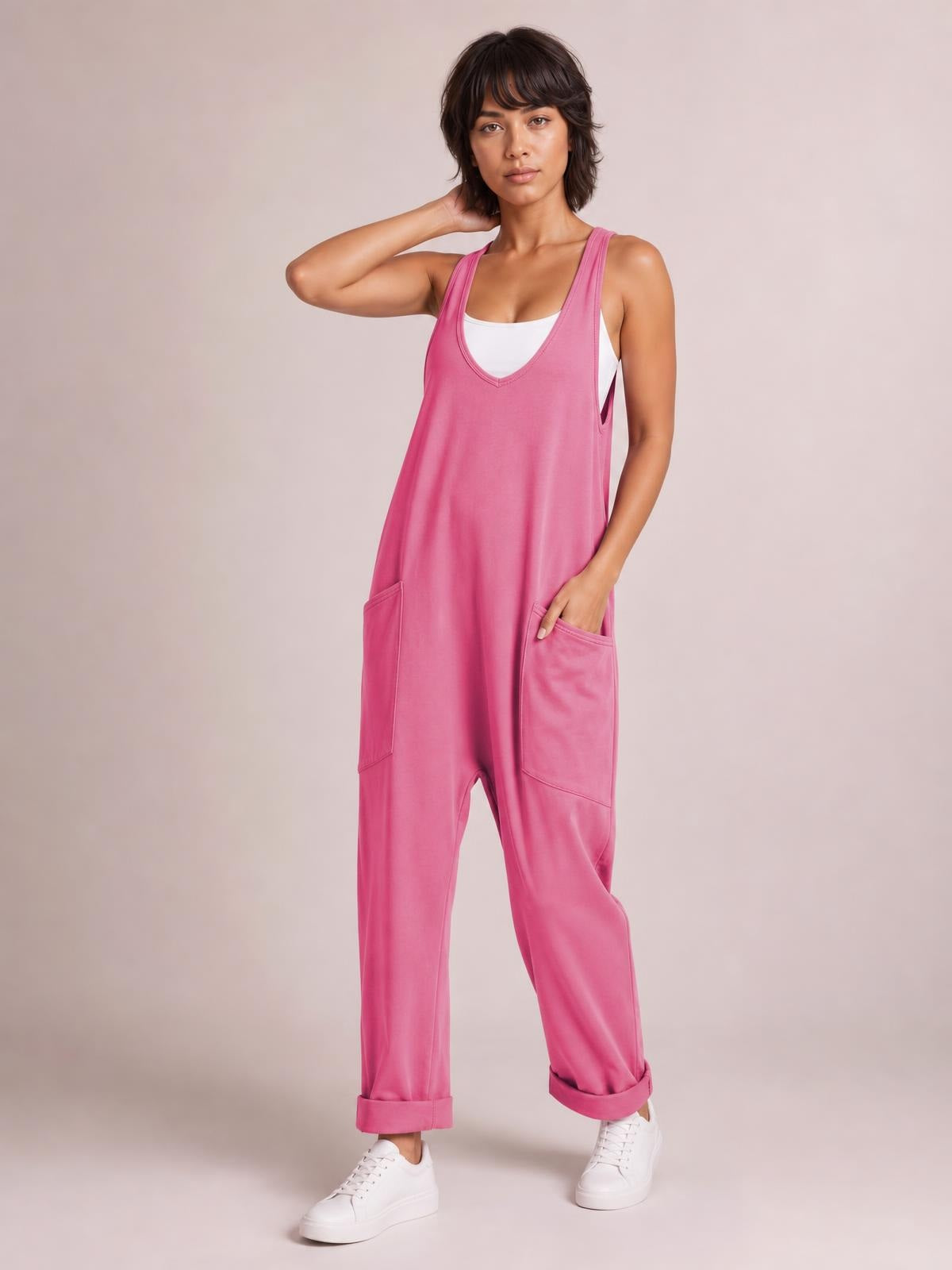 Model poses in a pink wide-strap jumpsuit with a V-neck, relaxed straight-leg silhouette, large patch pockets, and rolled ankle cuffs over a white tank.