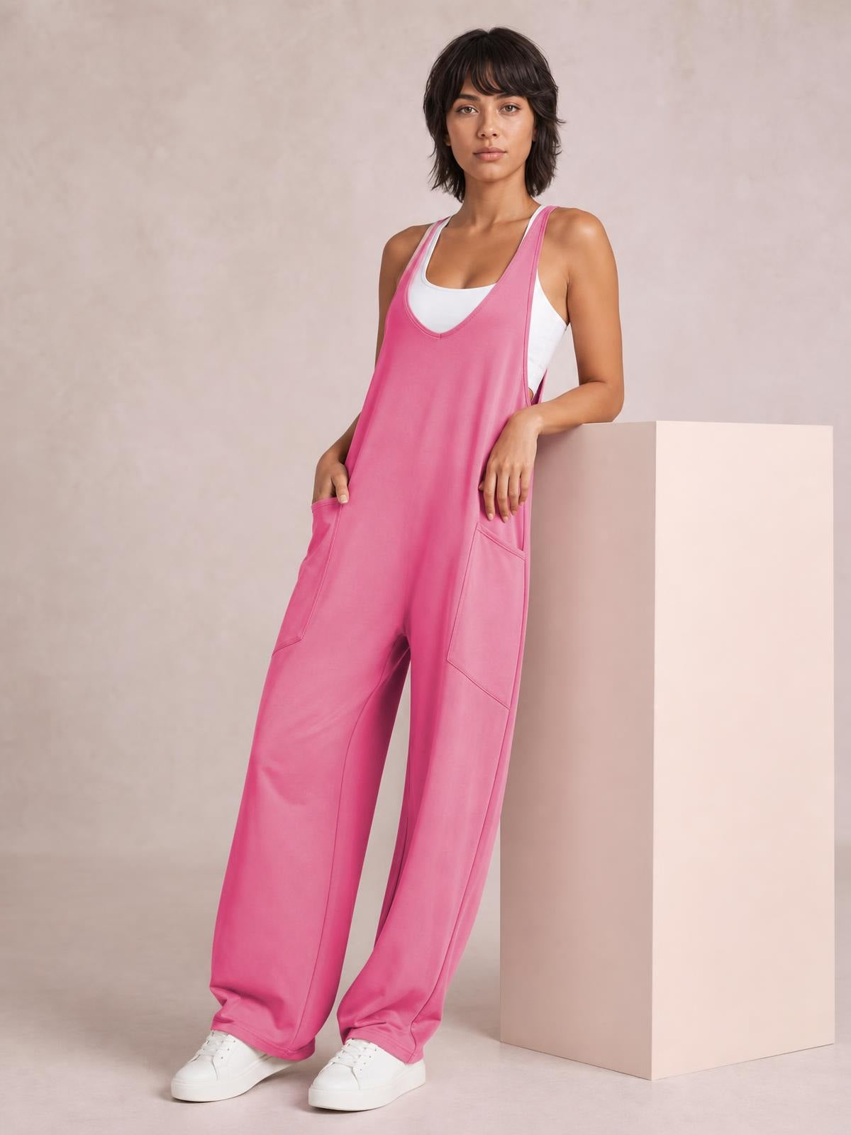 A model wears a pink wide-strap jumpsuit with a relaxed wide-leg silhouette, large front pockets, and a white tank layered underneath while leaning casually.