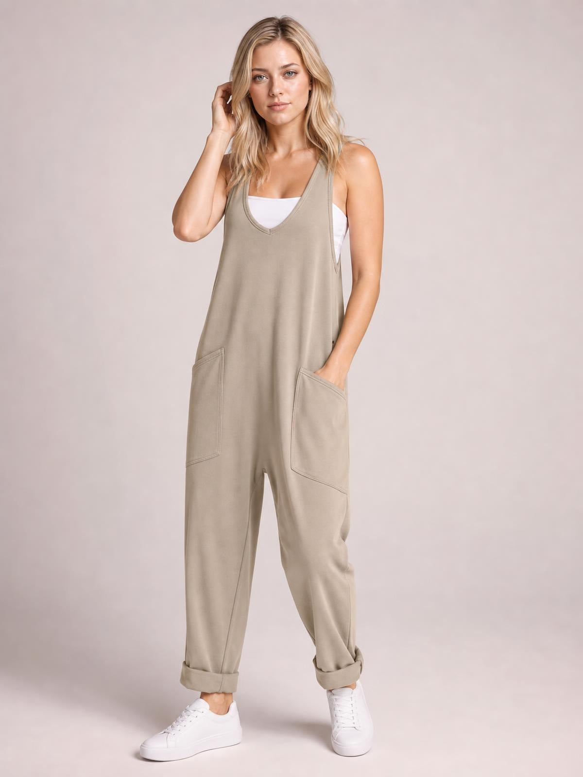 Model wearing a taupe wide-strap jumpsuit with a V-neck, relaxed tapered legs, rolled cuffs, and large front pockets, posing with one hand tucked inside.