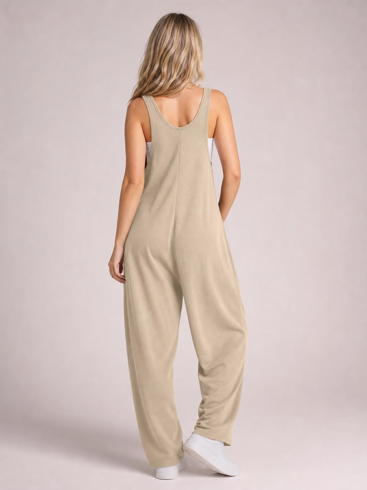Model stands with one hand in a pocket, showcasing a beige wide strap jumpsuit with a scoop back, center seam, and relaxed wide-leg silhouette.