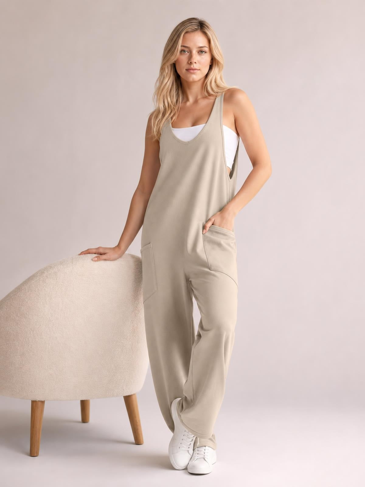 Model poses in a beige wide-strap jumpsuit with a relaxed straight-leg silhouette, scoop neckline, and front pockets, layered over a white top.