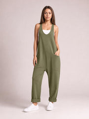 Model wearing an olive green wide-strap jumpsuit with a V-neck, relaxed tapered legs, large patch pockets, cuffed hems, and one hand tucked inside.
