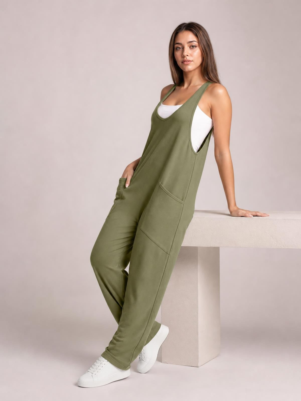 Model poses in an olive green wide strap jumpsuit with a relaxed straight-leg silhouette, V-neck bib, side pockets, and a white tank top underneath.