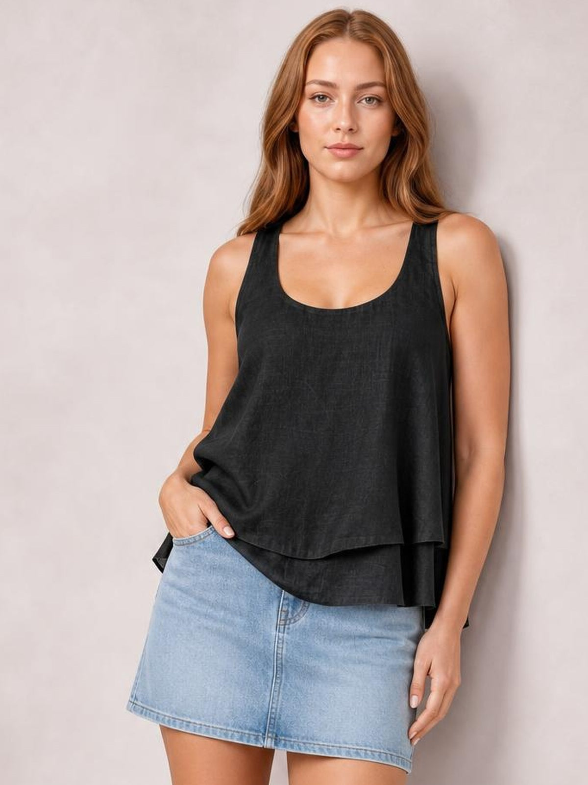 Model poses in a black wide-strap layered tank with a sheer, relaxed silhouette and scoop neckline, lifting the hem slightly over a denim skirt.